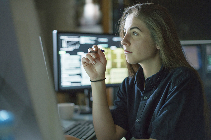 A young woman is seated at a desk surrounded by monitors displaying data